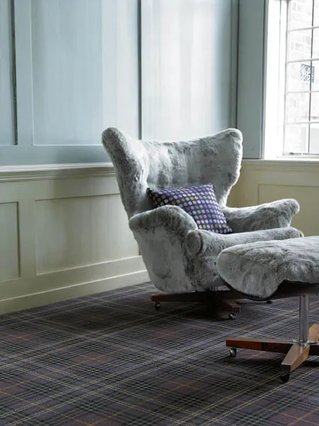 Purple and grey tartan carpet in a panelled room, with a grey sheepskin armchair and footstool.