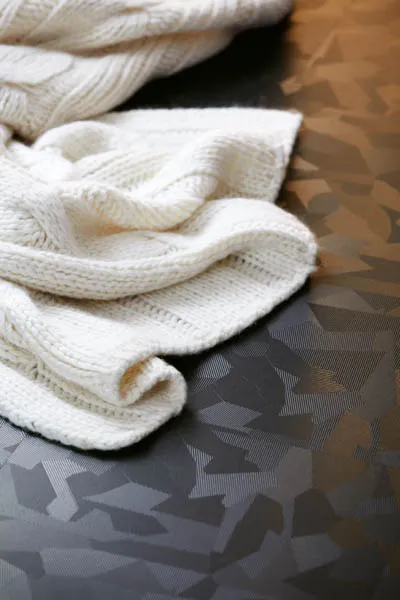 Cream cable-knit throw resting on a black laminate floor with a faceted geometric sheen.